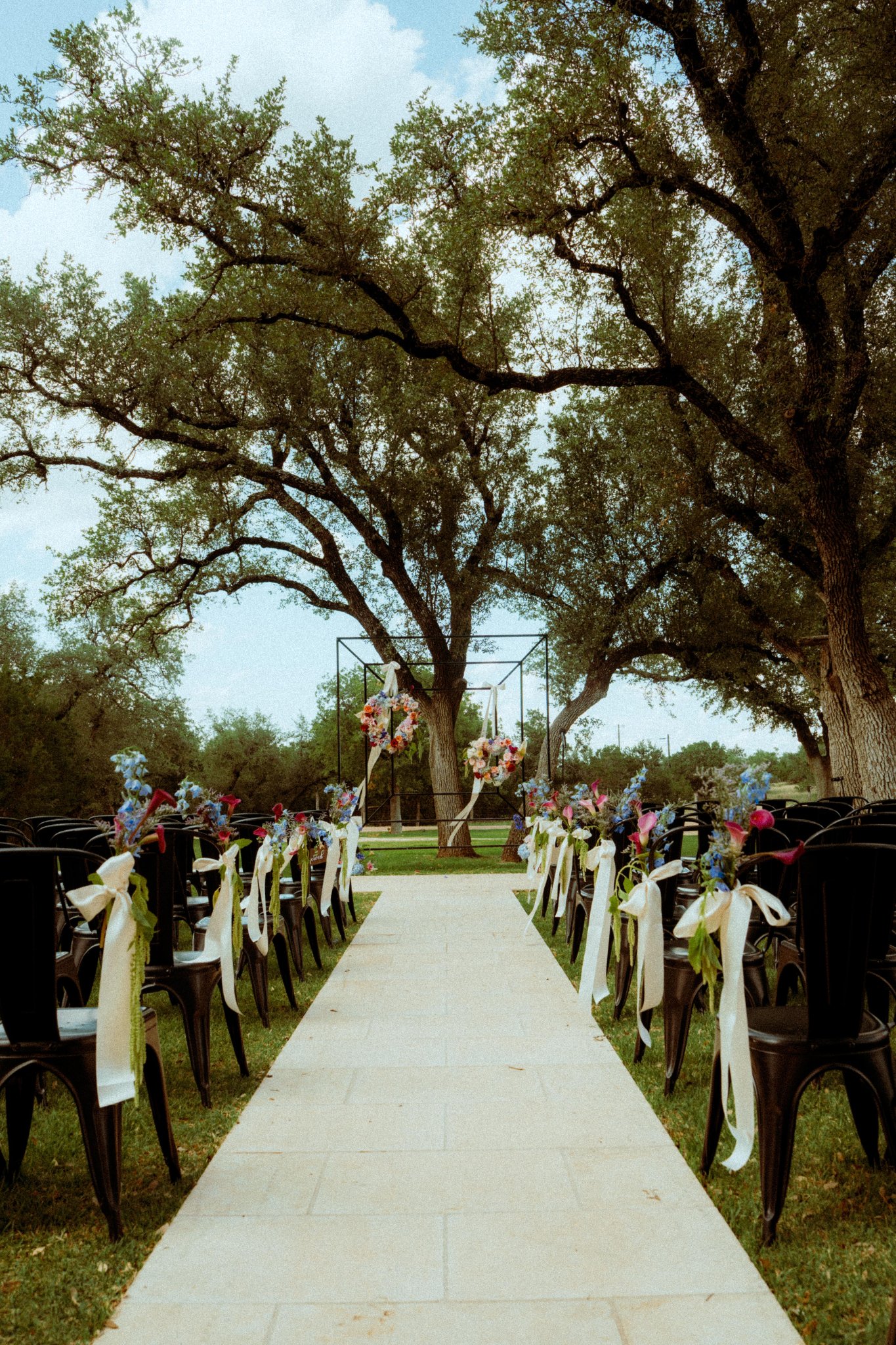 Bride and groom kissing at outdoor wedding ceremony at Willow Sky Ranch near Fredericksburg, TX — stone aisle with black farmhouse chairs and wildflower bouquets under sweeping live oak trees, Hill Country wedding venue near Austin and San Antonio, photographed by Zach Brigham Delude Photography